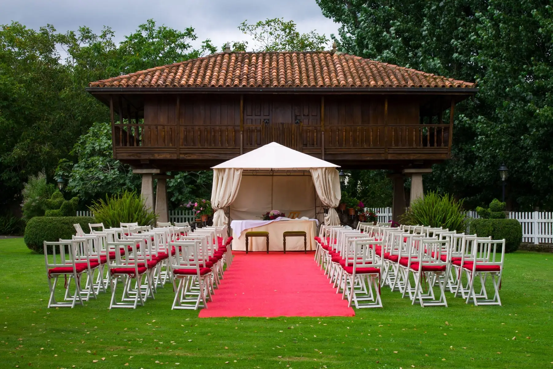 Sillas blancas con cojines rojos y una alfombra roja delante de un hórreo asturiano, con un pabellón blanco para la ceremonia